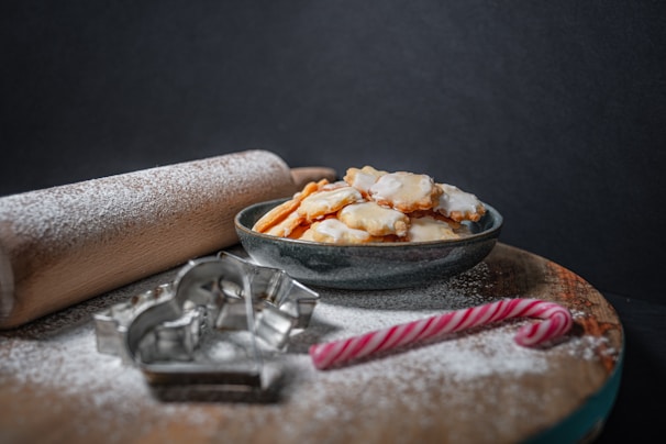  bowl filled with glazed cookies is set on a wooden board dusted with powdered sugar. Nearby, a wooden rolling pin, metal cookie cutters, and a candy cane are visible, all suggesting a baking theme.