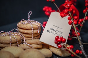 A stack of round cookies wrapped with red and white twine is placed next to a branch of vibrant red berries. A beige tag with 'Handgemacht in Santa's Werkstatt' printed on it is attached to the berries, adding a festive touch to the arrangement.
