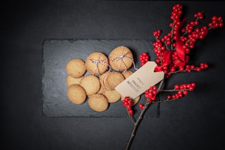 A collection of cookies tied with string is placed on a dark slate surface. Beside them is a sprig of red berries adorned with a red ribbon and a tag with writing.