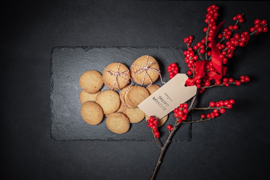 A collection of cookies tied with string is placed on a dark slate surface. Beside them is a sprig of red berries adorned with a red ribbon and a tag with writing.