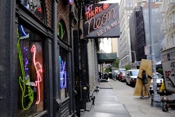 A city street with a storefront featuring several colorful neon signs including a flamingo and a guitar. The scene includes a neon sign displaying contact information. On the sidewalk, a person carries a large wooden board. The perspective shows a row of buildings extending into the distance on a slightly overcast day.