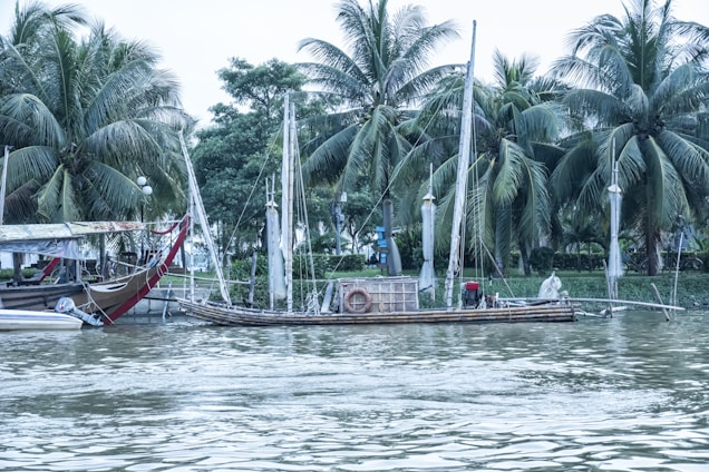 A tranquil riverside scene in Thailand at dawn, with soft light illuminating lush greenery and a traditional wooden boat.