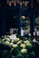 A market stall displays an assortment of fresh fruits and vegetables. Large green cabbages are prominent in the foreground, along with eggplants and root vegetables. Price tags with numbers are visible, indicating the cost of the produce. Hanging above the stall are bundles of garlic and dried goods, adding a rustic touch.