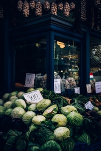 A market stall displays an assortment of fresh fruits and vegetables. Large green cabbages are prominent in the foreground, along with eggplants and root vegetables. Price tags with numbers are visible, indicating the cost of the produce. Hanging above the stall are bundles of garlic and dried goods, adding a rustic touch.