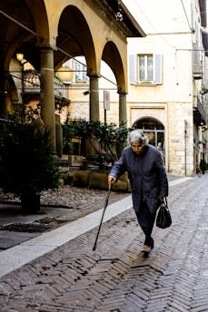 An elderly person with gray hair walks down a cobbled street using a cane for support. They wear a long, dark coat and hold a black handbag. The scene appears to be in an old European city with arched colonnades and rustic stone buildings. Potted plants and lanterns add to the ambiance.