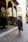 An elderly person with gray hair walks down a cobbled street using a cane for support. They wear a long, dark coat and hold a black handbag. The scene appears to be in an old European city with arched colonnades and rustic stone buildings. Potted plants and lanterns add to the ambiance.
