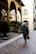 An elderly person with gray hair walks down a cobbled street using a cane for support. They wear a long, dark coat and hold a black handbag. The scene appears to be in an old European city with arched colonnades and rustic stone buildings. Potted plants and lanterns add to the ambiance.