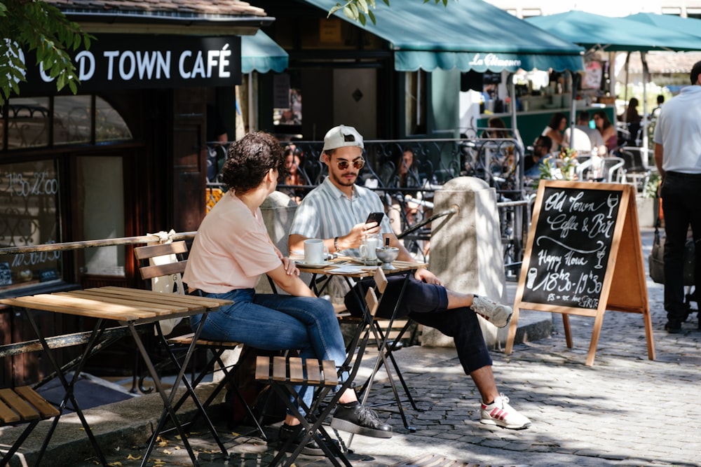 hombre y mujer sentados frente a una mesa plegable frente a una cafetería con toldos verdes