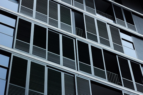 A building facade featuring numerous rectangular louvered windows arranged in a repetitive pattern. The structure is composed of sleek, modern lines and has a minimalist design, with metallic frames.