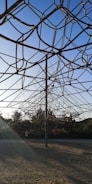 A large climbing structure made of ropes is situated in an outdoor play area, with a sandy ground and surrounded by greenery. The ropes form a geometric, web-like pattern connected to a central pole. The background shows trees under a clear blue sky, contributing to an inviting and serene atmosphere.