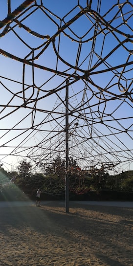 A large climbing structure made of ropes is situated in an outdoor play area, with a sandy ground and surrounded by greenery. The ropes form a geometric, web-like pattern connected to a central pole. The background shows trees under a clear blue sky, contributing to an inviting and serene atmosphere.