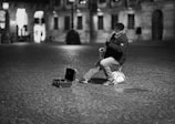 A street musician wearing a jacket and cap sits on a small chair playing a saxophone in an empty, cobblestone plaza. An open instrument case lies on the ground nearby. The background features historical buildings softly lit with a few people in the distance.