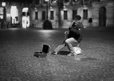 A street musician wearing a jacket and cap sits on a small chair playing a saxophone in an empty, cobblestone plaza. An open instrument case lies on the ground nearby. The background features historical buildings softly lit with a few people in the distance.