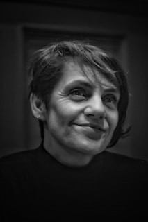 A black and white portrait of a person with short hair, wearing a dark turtleneck sweater. The person has a slight smile and is looking slightly off-camera. The background is blurred, focusing attention on the person's face.