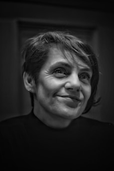 A black and white portrait of a person with short hair, wearing a dark turtleneck sweater. The person has a slight smile and is looking slightly off-camera. The background is blurred, focusing attention on the person's face.