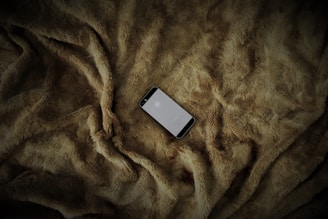 A smartphone is placed on a soft, textured, brown fur blanket. The phone screen is lit, displaying a logo.