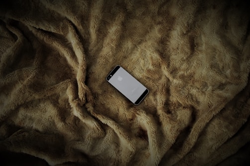 A smartphone is placed on a soft, textured, brown fur blanket. The phone screen is lit, displaying a logo.