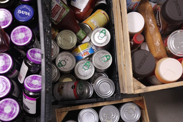 Various food items are stored in wooden crates. The assortment includes cans with green pull tabs, jars with purple lids, bottles, and boxes. The items are tightly packed, suggesting storage or organization.
