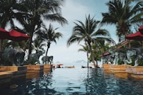 A luxurious infinity pool surrounded by palm trees and elephant statues overlooking a serene sea view. The scene includes red umbrellas and loungers with a backdrop of islands on the horizon. The atmosphere is tranquil and tropical.