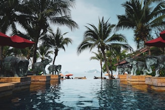 A luxurious infinity pool surrounded by palm trees and elephant statues overlooking a serene sea view. The scene includes red umbrellas and loungers with a backdrop of islands on the horizon. The atmosphere is tranquil and tropical.