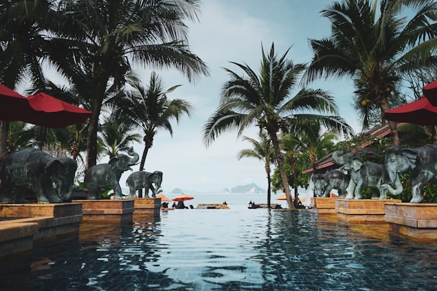 A luxurious infinity pool surrounded by palm trees and elephant statues overlooking a serene sea view. The scene includes red umbrellas and loungers with a backdrop of islands on the horizon. The atmosphere is tranquil and tropical.