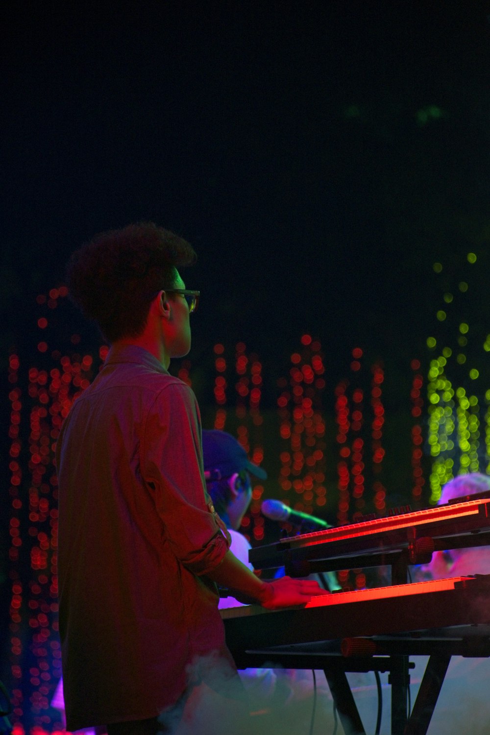 赤い長袖シャツを着て眼鏡をかけ、ピアノを弾きながら立っている男性