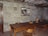 A rustic room with a large wooden table surrounded by wooden chairs. The wall is made of stone, and there is a framed black-and-white photo hanging on it. Several wooden bowls and a colorful ceramic vase are placed on the table. The ceiling features exposed wooden beams.