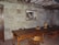 A rustic room with a large wooden table surrounded by wooden chairs. The wall is made of stone, and there is a framed black-and-white photo hanging on it. Several wooden bowls and a colorful ceramic vase are placed on the table. The ceiling features exposed wooden beams.