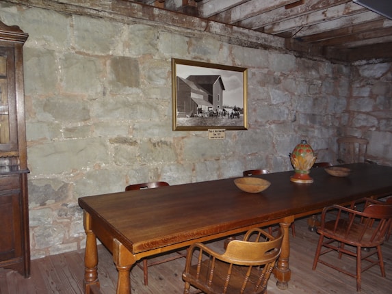 A rustic room with a large wooden table surrounded by wooden chairs. The wall is made of stone, and there is a framed black-and-white photo hanging on it. Several wooden bowls and a colorful ceramic vase are placed on the table. The ceiling features exposed wooden beams.