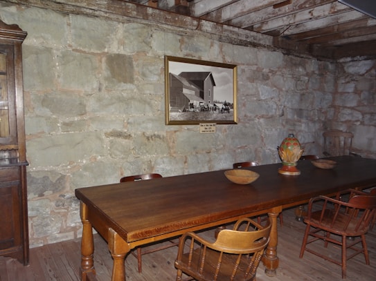 A rustic room with a large wooden table surrounded by wooden chairs. The wall is made of stone, and there is a framed black-and-white photo hanging on it. Several wooden bowls and a colorful ceramic vase are placed on the table. The ceiling features exposed wooden beams.
