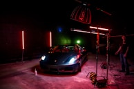 A sleek sports car is illuminated under dramatic lighting in a dimly lit garage or studio setting. The lighting includes red and green neon effects with the car prominently positioned. Photography equipment and a person stand nearby, adding to the setup's professional appearance.