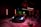 A sleek sports car is illuminated under dramatic lighting in a dimly lit garage or studio setting. The lighting includes red and green neon effects with the car prominently positioned. Photography equipment and a person stand nearby, adding to the setup's professional appearance.