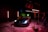 A sleek sports car is illuminated under dramatic lighting in a dimly lit garage or studio setting. The lighting includes red and green neon effects with the car prominently positioned. Photography equipment and a person stand nearby, adding to the setup's professional appearance.