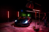 A sleek sports car is illuminated under dramatic lighting in a dimly lit garage or studio setting. The lighting includes red and green neon effects with the car prominently positioned. Photography equipment and a person stand nearby, adding to the setup's professional appearance.
