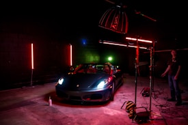 A sleek sports car is illuminated under dramatic lighting in a dimly lit garage or studio setting. The lighting includes red and green neon effects with the car prominently positioned. Photography equipment and a person stand nearby, adding to the setup's professional appearance.