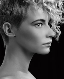 A black and white profile portrait of a person with short curly hair, sharp facial features, and a focused expression. The lighting highlights the texture of the hair and smoothness of the skin against a dark background.