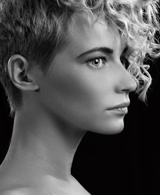 A black and white profile portrait of a person with short curly hair, sharp facial features, and a focused expression. The lighting highlights the texture of the hair and smoothness of the skin against a dark background.