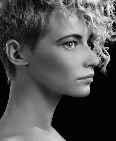 A black and white profile portrait of a person with short curly hair, sharp facial features, and a focused expression. The lighting highlights the texture of the hair and smoothness of the skin against a dark background.