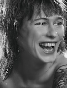 A person is laughing heartily with visible teeth. The image is in black and white, highlighting the contrast between light and shadow. The person has shaggy hair and a tattoo on their shoulder.