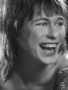 A person is laughing heartily with visible teeth. The image is in black and white, highlighting the contrast between light and shadow. The person has shaggy hair and a tattoo on their shoulder.