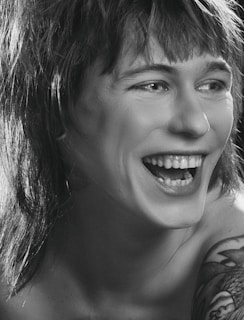 A person is laughing heartily with visible teeth. The image is in black and white, highlighting the contrast between light and shadow. The person has shaggy hair and a tattoo on their shoulder.