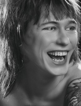 A person is laughing heartily with visible teeth. The image is in black and white, highlighting the contrast between light and shadow. The person has shaggy hair and a tattoo on their shoulder.