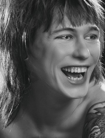 A person is laughing heartily with visible teeth. The image is in black and white, highlighting the contrast between light and shadow. The person has shaggy hair and a tattoo on their shoulder.