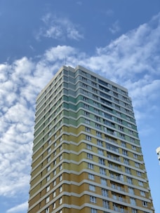 A tall multi-story building with a yellow and green exterior stands under a blue sky with scattered clouds. The building has numerous windows and appears to be residential or commercial.