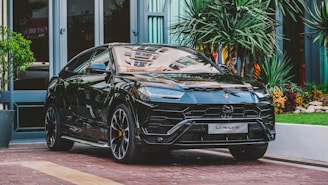 A sleek black luxury SUV is parked on a brick driveway surrounded by lush greenery and modern architecture. The vehicle boasts a polished exterior with detailed design elements and sports large alloy wheels.