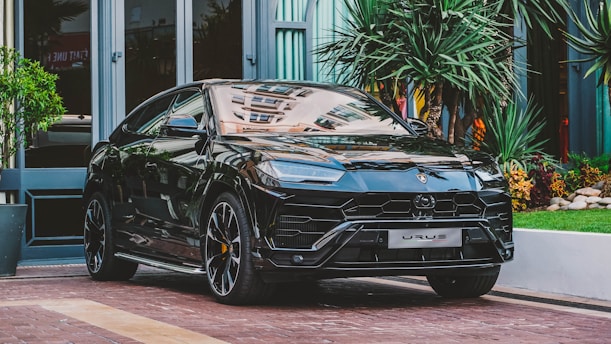 A sleek black luxury SUV is parked on a brick driveway surrounded by lush greenery and modern architecture. The vehicle boasts a polished exterior with detailed design elements and sports large alloy wheels.