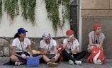 Four young women are sitting on a sidewalk against a wall covered with climbing plants. They are casually dressed, wearing hats or headscarves, and lanyards around their necks. They appear to be talking and relaxing, with bags and belongings around them.