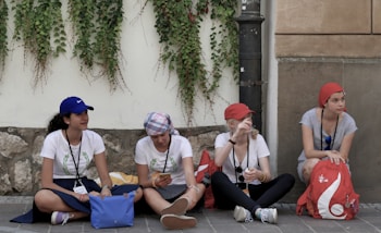 Four young women are sitting on a sidewalk against a wall covered with climbing plants. They are casually dressed, wearing hats or headscarves, and lanyards around their necks. They appear to be talking and relaxing, with bags and belongings around them.