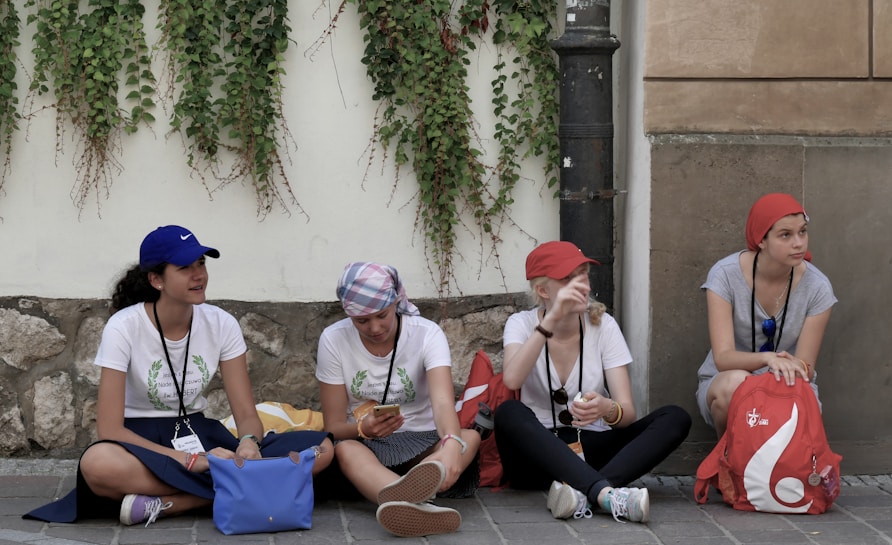 Four young women are sitting on a sidewalk against a wall covered with climbing plants. They are casually dressed, wearing hats or headscarves, and lanyards around their necks. They appear to be talking and relaxing, with bags and belongings around them.