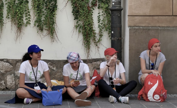 Four young women are sitting on a sidewalk against a wall covered with climbing plants. They are casually dressed, wearing hats or headscarves, and lanyards around their necks. They appear to be talking and relaxing, with bags and belongings around them.